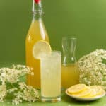 Elderflower syrup in a tall bottle and a refreshing elderflower drink in a glass. Lemon slices and elderflower heads are next to the glass.