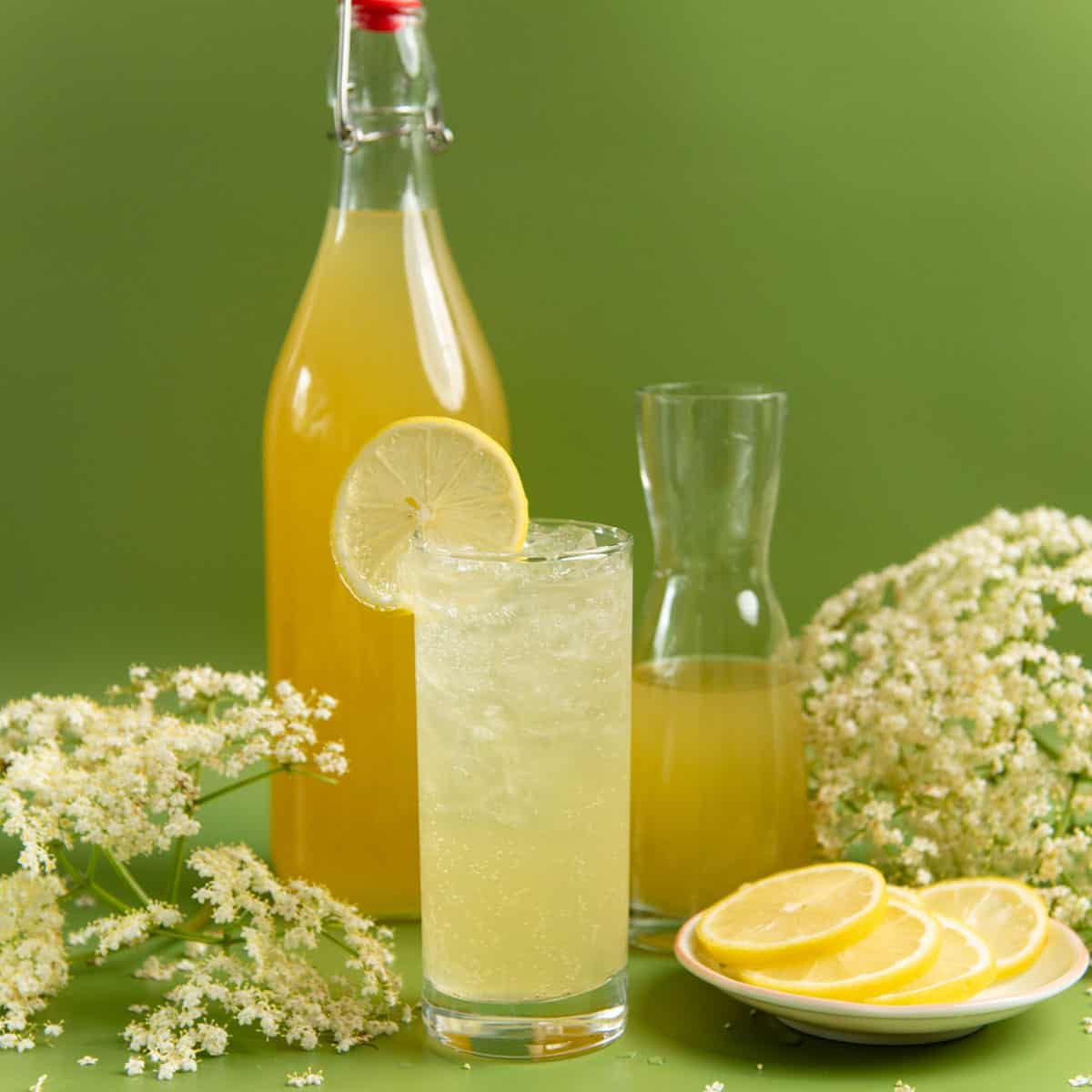 Elderflower syrup in a tall bottle and a refreshing elderflower drink in a glass. Lemon slices and elderflower heads are next to the glass.
