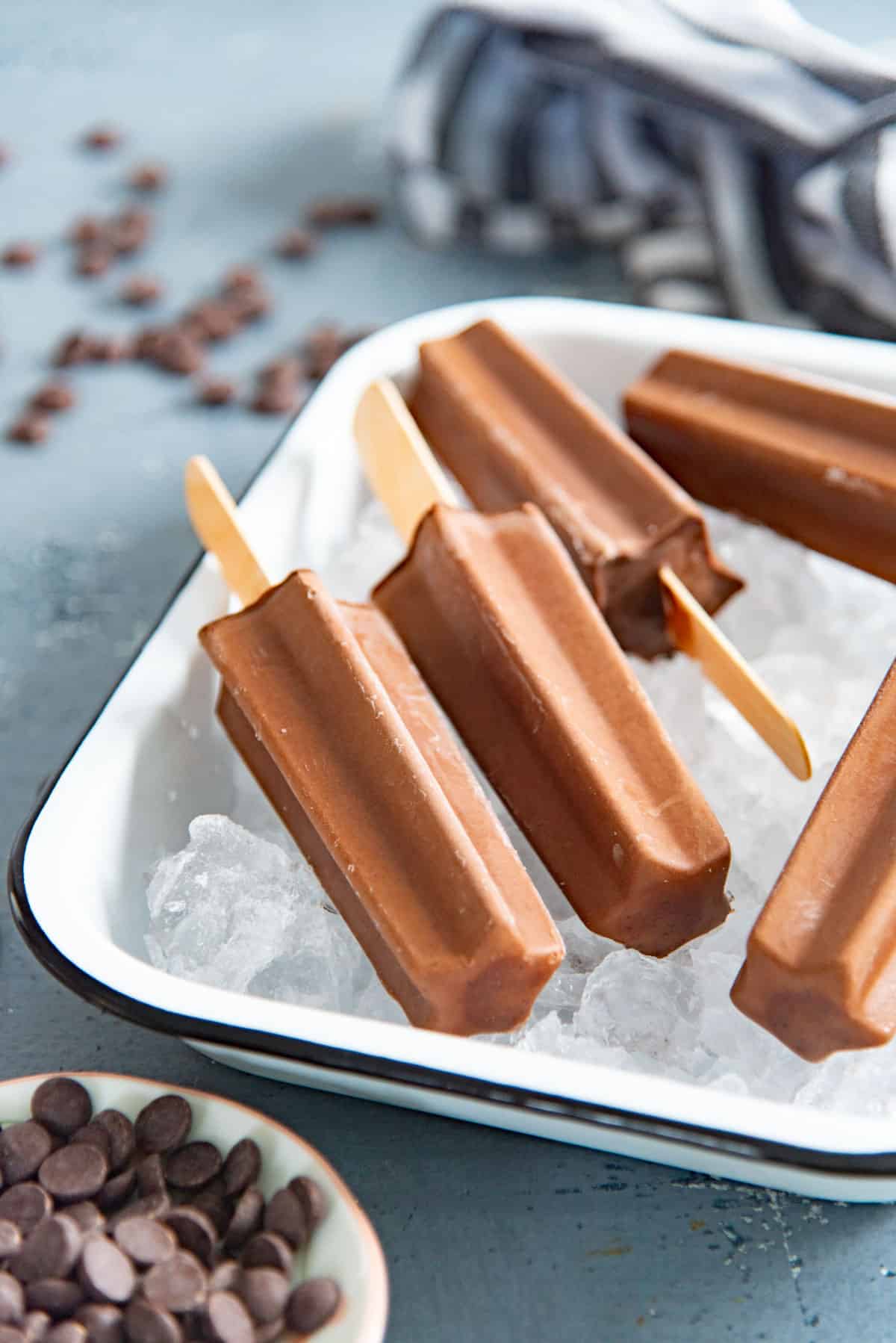 Several chocolate fudgesicles placed on a bed of ice in a white dish. They look creamy and rich.