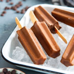 Several chocolate fudgesicles placed on a bed of ice in a white dish. Chocolate chips are in a small plate are in the foreground.