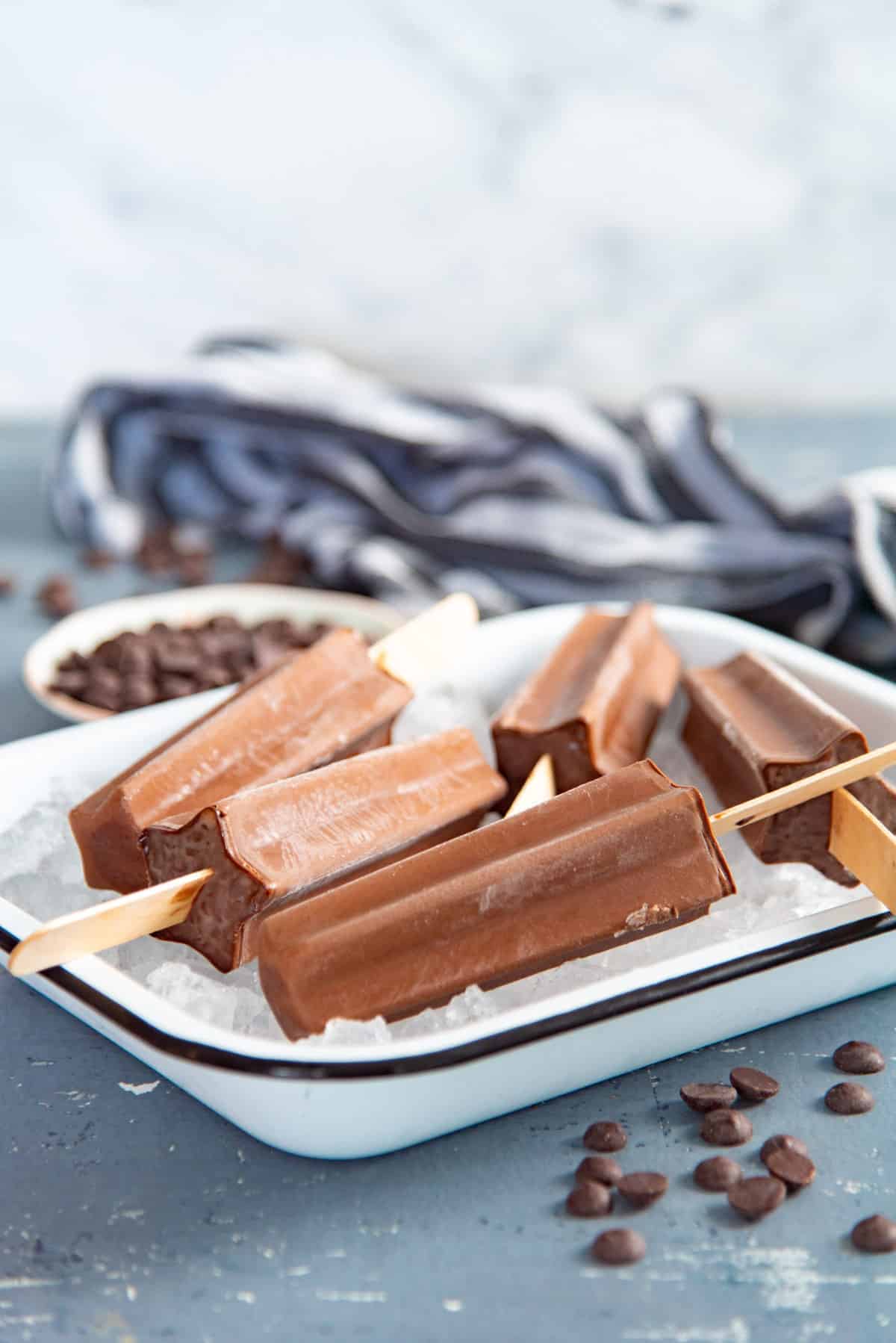 Several creamy fudgesicles placed on top of crushed ice in a white dish. There are chocolate chips scattered around the dish.