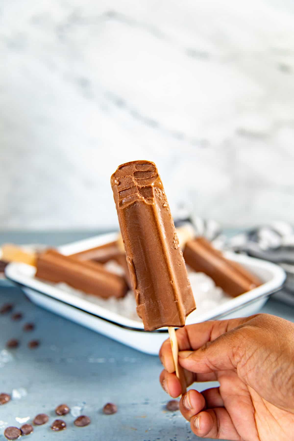 A hand holding a chocolate popsicle with a bite taken out of it at the top. More popsicles in the background.