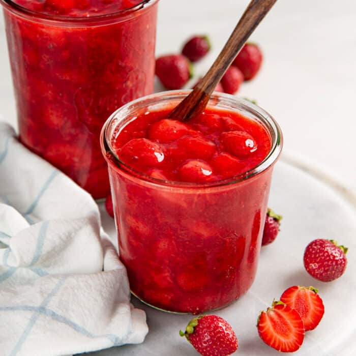 Close up of the thick, red, shiny strawberry sauce topping in a glass jar with a wooden spoon.