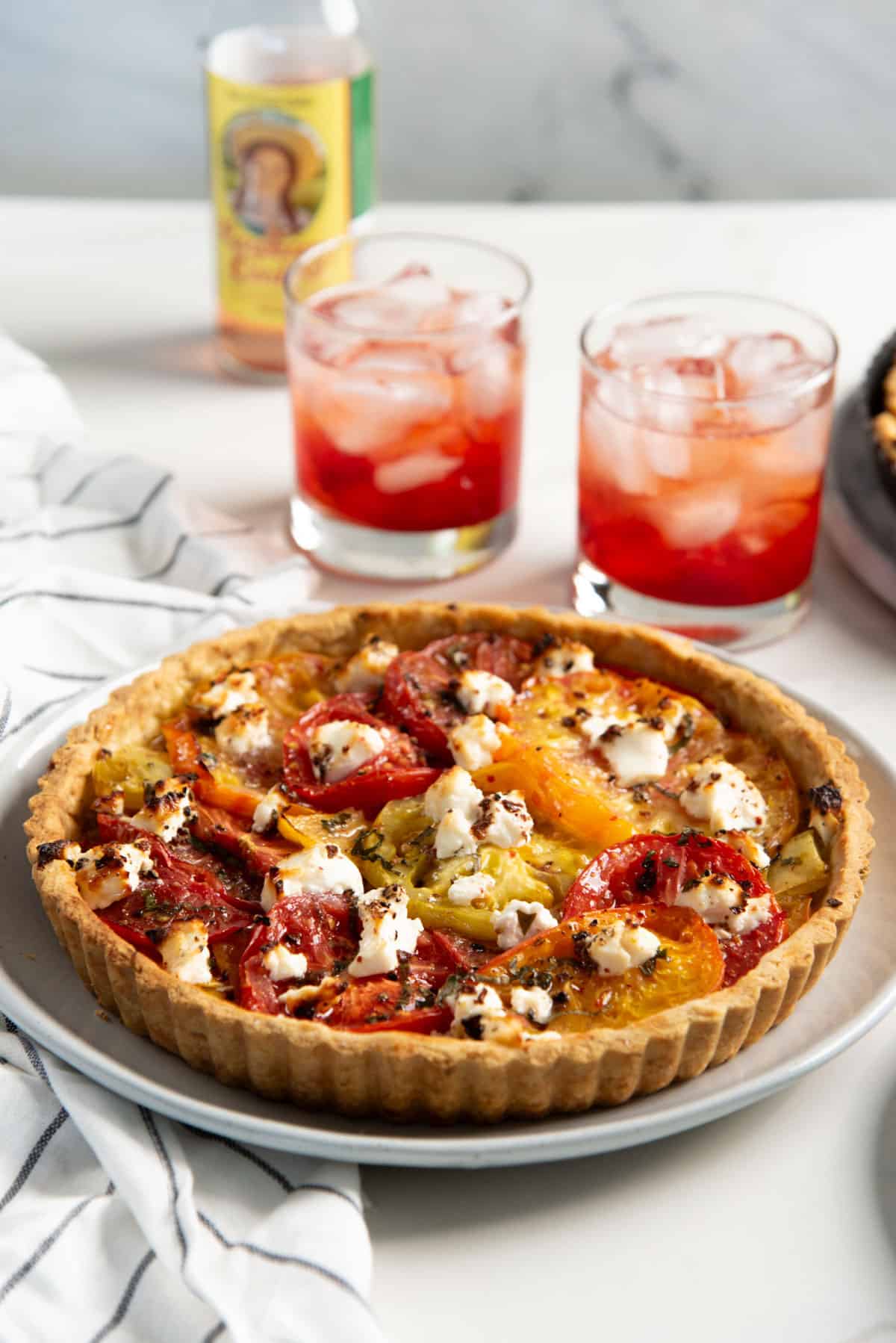 Freshly baked fresh tomato tart in a tart pan served on a white plate, and two glasses with a red colored drink next to it.