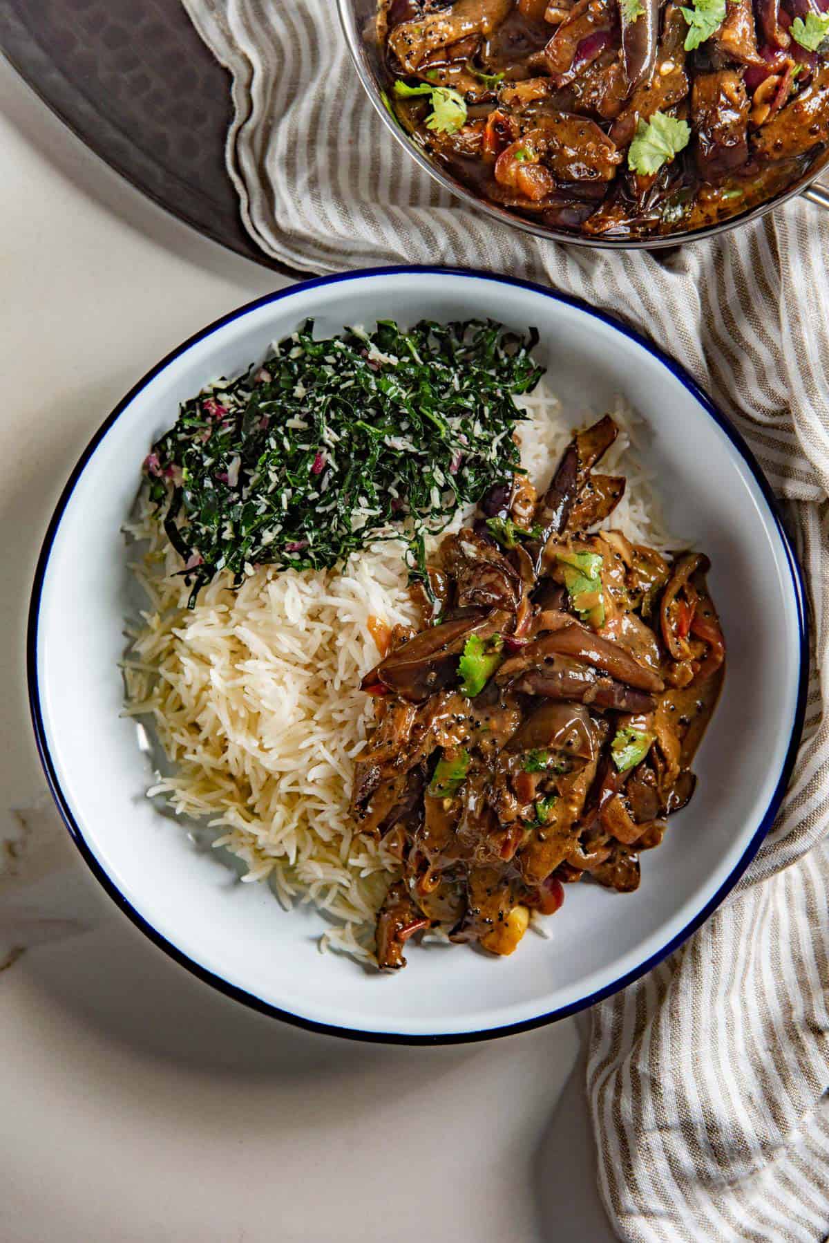 Overhead view of eggplant curry served in a white plate with steamed white rice and a leafy green side dish.