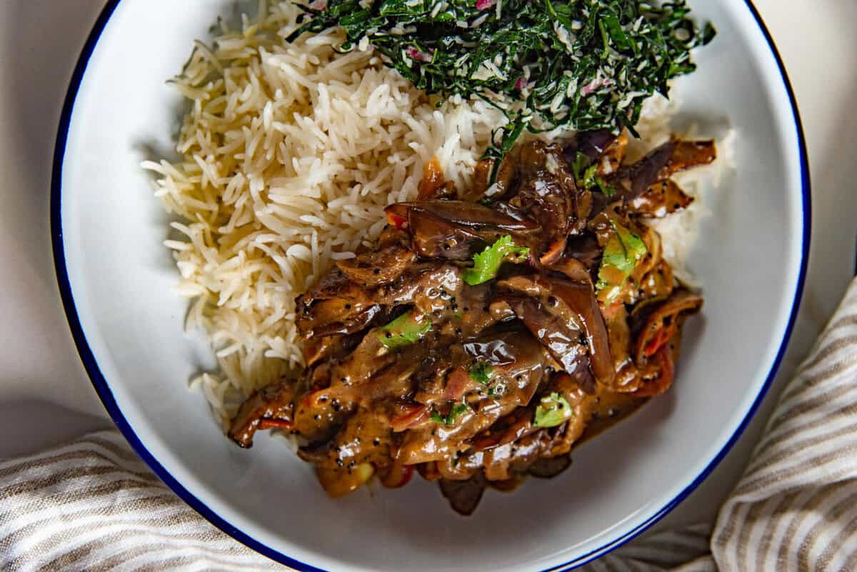 Close up of brinjal curry served in a white plate with white rice and a leafy green salad.