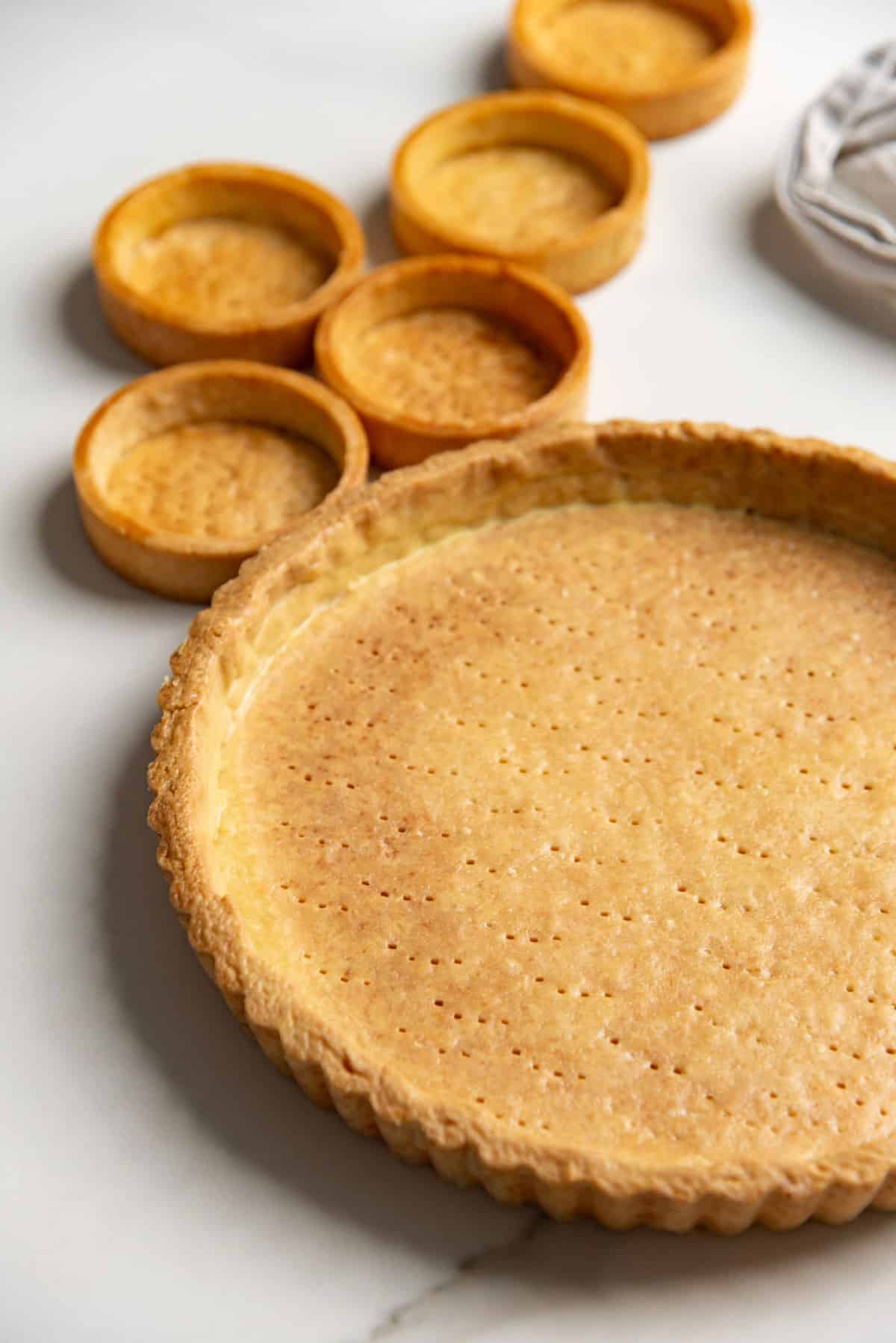 Close up image of a blind baked large tart and several mini tarts using shortcrust pastry dough. The crusts look golden brown.