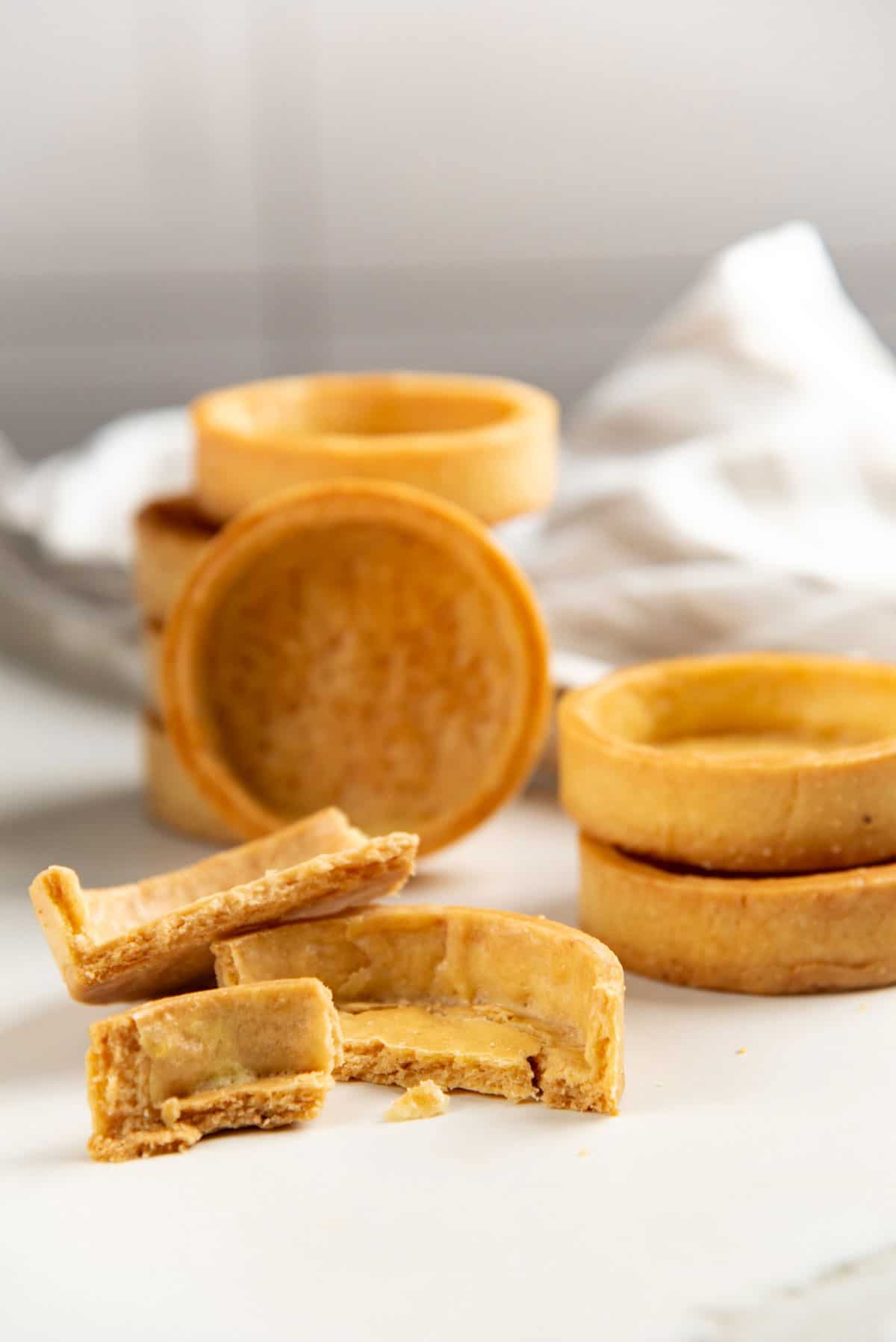 A mini pate brisee tart crust broken into several pieces to show the crumbly texture. More in tact tart crusts can be seen in the background.