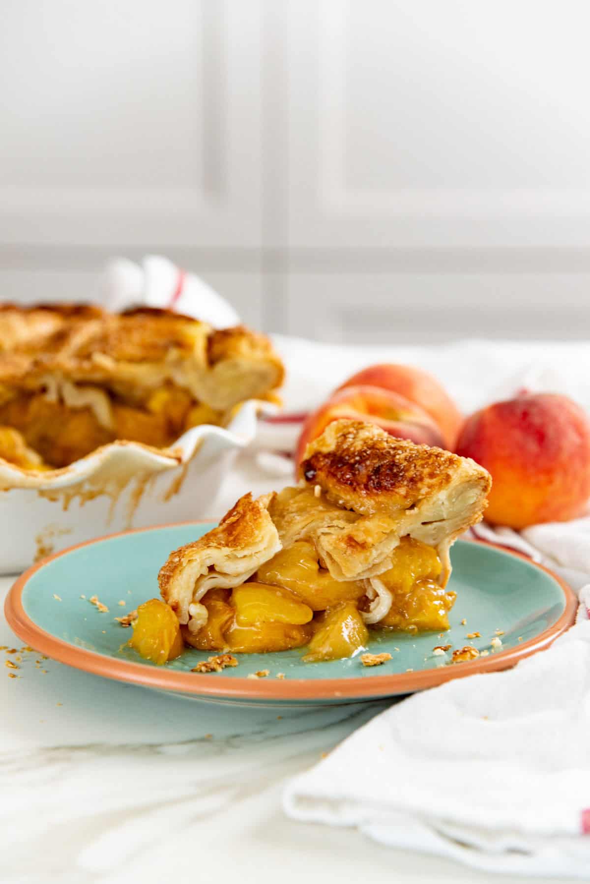 Fresh peach pie slice served on a small green plate. Fresh whole peaches and a deep pie dish with a peach pie in the background.