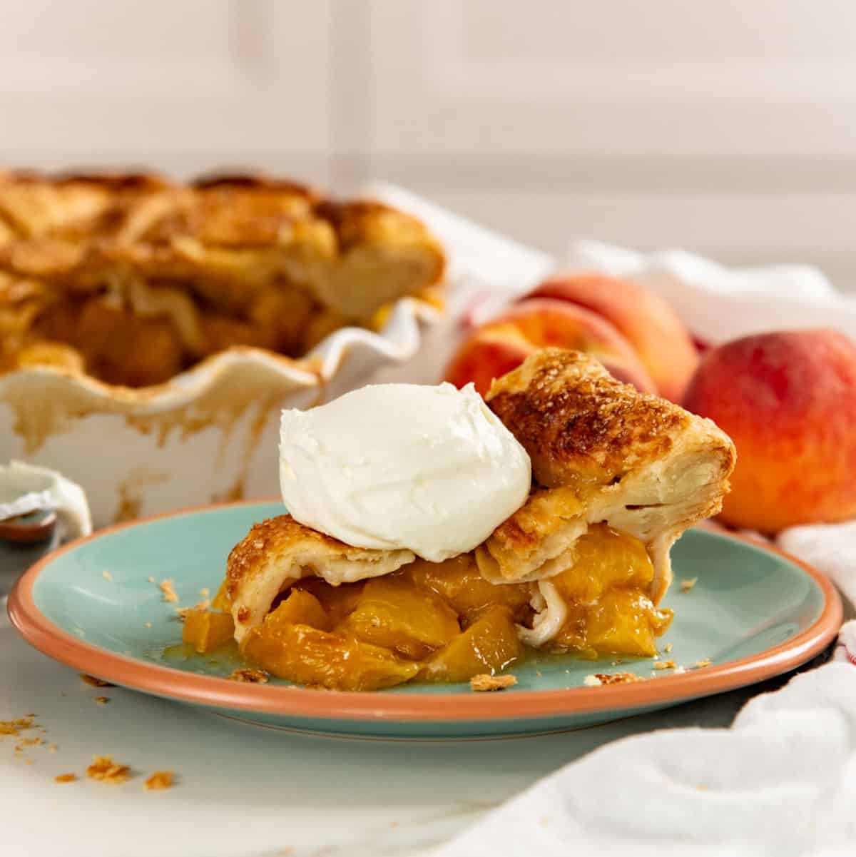 Slice of peach pie served with whipped cream on a small plate. Rest of the pie and fresh peaches seen in the background.